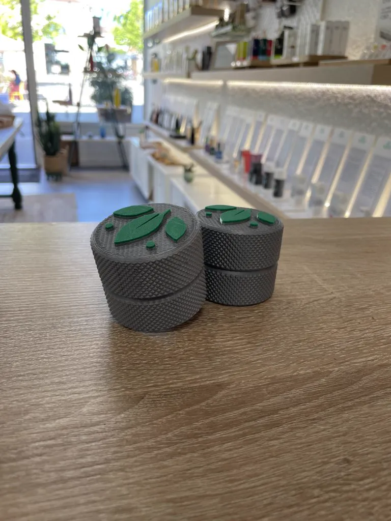 Deux petites boîtes moletées grises imprimées en 3D, au couvercle orné d’un logo vert en relief, posées sur un comptoir