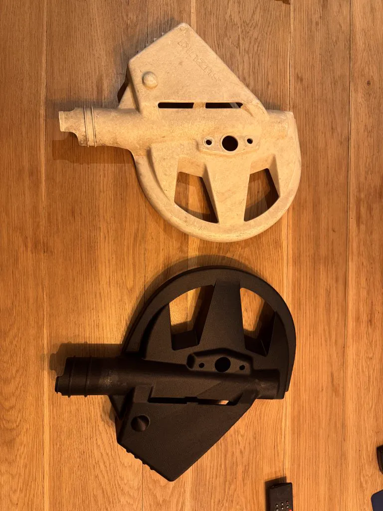 Une pièce d’origine claire, usée, et sa copie imprimée en 3D, noire, posées sur un plan de travail en bois